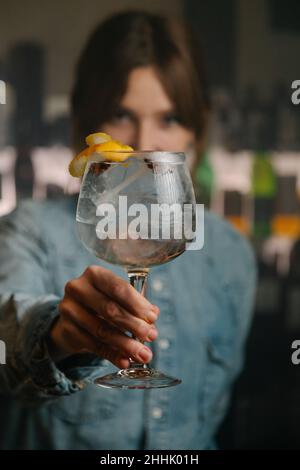 Die Barkeeperin mit weichem Fokus blickt auf die Kamera, während sie Weinglas mit Gin und Tonic-Cocktail serviert, der mit orangefarbener Schale auf der Holztheke in der Bar dekoriert ist Stockfoto