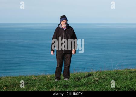 Ganzer Körper eines positiven älteren bärtigen Mannes in Oberbekleidung, der auf einem grasbewachsenen Hügel an der Küste des blauen plätschernden Meeres steht und die Kamera anschaut Stockfoto