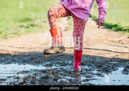 Crop unkenntlich Kind in rosa Hosen und roten Gummistiefel stehen auf schmutzigen Weg mit Pfützen auf der Straße der Stadt Stockfoto
