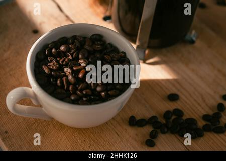 Von oben auf Haufen frisch gerösteten Kaffeebohnen in weißer Keramikschale serviert auf Holztisch in der Nähe von Französisch Presse Stockfoto