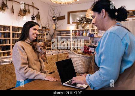Seitenansicht einer ruhigen Verkäuferin in legerer Kleidung, die auf einem Laptop mit schwarzem Bildschirm steht und tippt, während sie vor positiv an der Holztheke steht Stockfoto