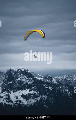 Anonyme Person, die mit einem bunten Gleitschirm über verschneiten Bergen fliegt, die von Nadelwäldern bedeckt sind, unter trübem, düsteren Himmel Stockfoto