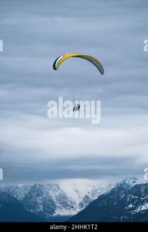 Anonyme Person, die mit einem bunten Gleitschirm über verschneiten Bergen fliegt, die von Nadelwäldern bedeckt sind, unter trübem, düsteren Himmel Stockfoto