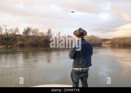 Rückansicht eines nicht erkennbaren bärtigen männlichen Wanderers in warmer Kleidung, der moderne Drohnen fliegt, während er während des Trekkings bei Sonnenuntergang in der Nähe des Sees steht Stockfoto