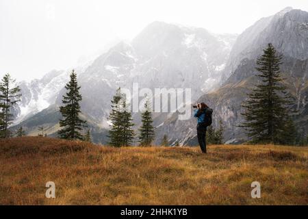 Seitenansicht eines anonymen männlichen Touristen mit Rucksack, der auf einer Wiese in Österreich auf verschneiten Hochbergen fotografiert Stockfoto