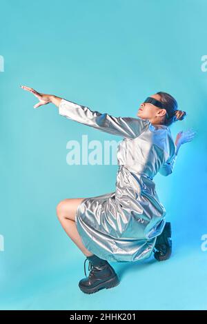 Seitenansicht einer modischen jungen ethnischen Frau in futuristischem Outfit und einer VR-Brille, die auf Geisterfeldern sitzt und gestikiert, während sie in die virtuelle Realität eingeht Stockfoto