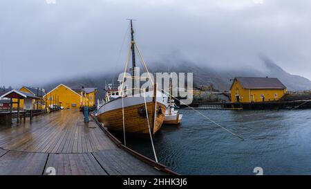 Große Fischerboote, die im Meer in der Nähe eines hölzernen Piers in einem kleinen Dorf in Nusfjord festgemacht sind, umgeben von felsigen Bergen vor nebligen Himmel Stockfoto