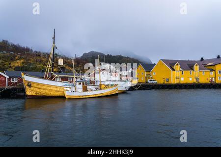 Große Fischerboote, die im Meer in der Nähe eines hölzernen Piers in einem kleinen Dorf in Nusfjord festgemacht sind, umgeben von felsigen Bergen vor nebligen Himmel Stockfoto