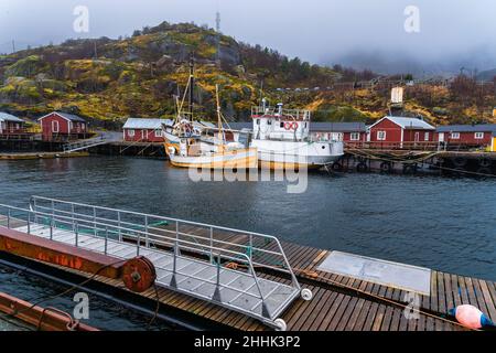 Große Fischerboote, die im Meer in der Nähe eines hölzernen Piers in einem kleinen Dorf in Nusfjord festgemacht sind, umgeben von felsigen Bergen vor nebligen Himmel Stockfoto