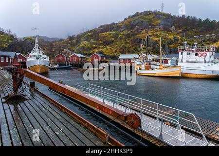 Große Fischerboote, die im Meer in der Nähe eines hölzernen Piers in einem kleinen Dorf in Nusfjord festgemacht sind, umgeben von felsigen Bergen vor nebligen Himmel Stockfoto