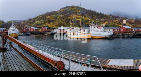 Große Fischerboote, die im Meer in der Nähe eines hölzernen Piers in einem kleinen Dorf in Nusfjord festgemacht sind, umgeben von felsigen Bergen vor nebligen Himmel Stockfoto
