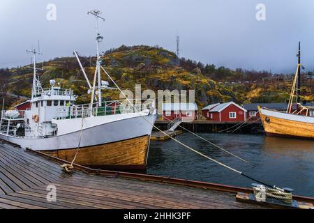 Große Fischerboote, die im Meer in der Nähe eines hölzernen Piers in einem kleinen Dorf in Nusfjord festgemacht sind, umgeben von felsigen Bergen vor nebligen Himmel Stockfoto