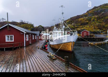 Große Fischerboote, die im Meer in der Nähe eines hölzernen Piers in einem kleinen Dorf in Nusfjord festgemacht sind, umgeben von felsigen Bergen vor nebligen Himmel Stockfoto