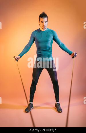 In voller Länge junger spanischer männlicher Athlet mit dunklem Haar in Sportswear, der mit einem Widerstandsseil vor orangefarbenem Hintergrund i trainiert Stockfoto