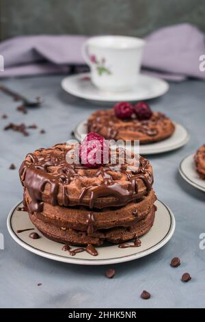 Leckere süße Waffeln mit Schokoladensauce und Himbeere serviert auf dem Tisch mit weißer Tasse und Schokoladentropfen in der hellen Küche Stockfoto