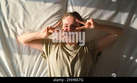Draufsicht auf eine fröhliche junge Dame, die lächelt und die Kamera anschaut, während sie am sonnigen Tag auf einem bequemen Bett mit erhobenem Arm liegt Stockfoto