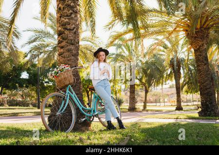 Stilvolle junge Frau in legerer Kleidung und Hut auf dem Fahrrad mit Blumen in der Nähe von Palmen und Surfen in sozialen Medien auf dem Smartphone an sonnigen Tag im Park Stockfoto
