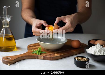 Crop gesichtslosen männlichen Koch zerbrechen rohes Ei in Schüssel auf Schneidebrett beim Kochen am Tisch mit verschiedenen Zutaten in der Küche Stockfoto