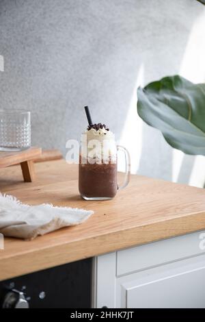 Glas mit Schokoladenfrappe und süßer Schlagsahne, serviert mit Stroh, auf Holztisch im hellen Raum Stockfoto