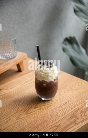 Glas mit Schokoladenfrappe und süßer Schlagsahne, serviert mit Stroh, auf Holztisch im hellen Raum Stockfoto