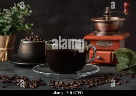 Ein Glas frisch gebrühten aromatischen Kaffees auf einer Untertasse, die mit verstreuten Bohnen gegen das manuelle Mahlwerk gelegt wird Stockfoto