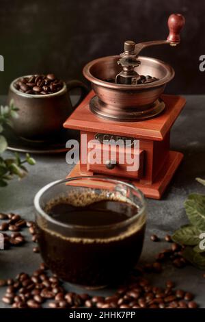 Ein Glas frisch gebrühten aromatischen Kaffees auf einer Untertasse, die mit verstreuten Bohnen gegen das manuelle Mahlwerk gelegt wird Stockfoto