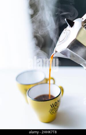 Frisch gebrühter, dampfender, heißer Kaffee, der aus einem Mokakatopf aus Metall in eine gelbe Tasse auf dem Tisch in der hellen Küche auf unscharfem Hintergrund gegossen wird Stockfoto