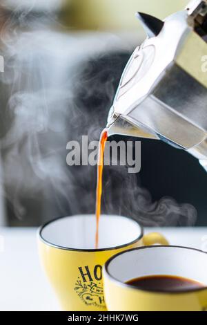 Frisch gebrühter, dampfender, heißer Kaffee, der aus einem Mokakatopf aus Metall in eine gelbe Tasse auf dem Tisch in der hellen Küche auf unscharfem Hintergrund gegossen wird Stockfoto
