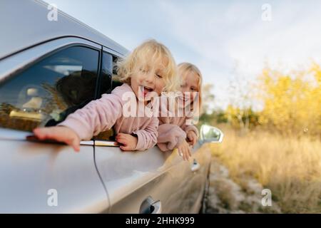 Niedliche Schwestern in legerer Kleidung, die aus dem Fenster schauen, während sie auf einer gemeinsamen Fahrt im Auto auf dem Land mit grünen Pflanzen sitzen Stockfoto