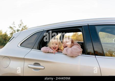 Niedliche Schwestern in legerer Kleidung, die aus dem Fenster schauen, während sie auf einer gemeinsamen Fahrt im Auto auf dem Land mit grünen Pflanzen sitzen Stockfoto