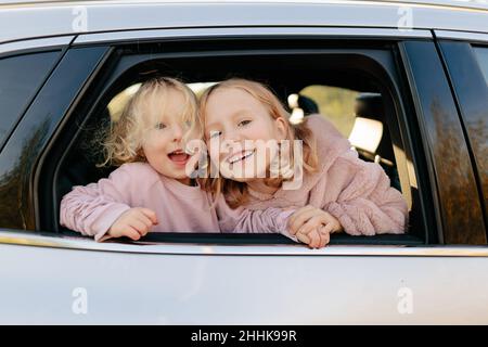 Niedliche Schwestern in legerer Kleidung, die aus dem Fenster schauen, während sie auf einer gemeinsamen Fahrt im Auto auf dem Land mit grünen Pflanzen sitzen Stockfoto