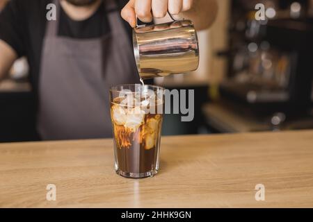 Schneiden Sie den männlichen Barista in der Schürze, und fügen Sie während des Arbeitstages Milch in Glas mit Eiskaffee im Kaffeehaus hinzu Stockfoto