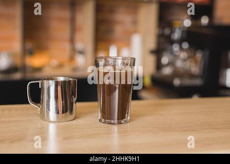 Glastasse mit aromatischem, vereisten Americano auf dem Bartisch in der Nähe eines Metallkänzers in einem hellen Café Stockfoto
