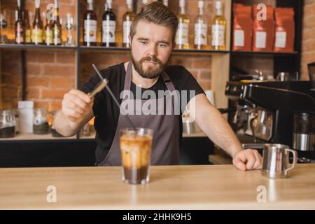 Bärtiger Barista in der Schürze, der Plastikstroh in ein Glas Eiskaffee mit Milch mit Zangen während der Arbeit im Café-Geschäft legte Stockfoto