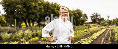 Fröhliche junge Köchin lächelt fröhlich auf einem landwirtschaftlichen Feld an der Kamera. Autarke Köchin, die mit gekreuzten Armen in einem steht Stockfoto