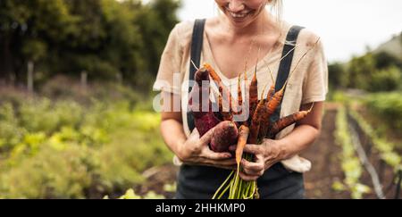 Fröhliche Bäuerin mit frisch gepflückten Karotten und Süßkartoffeln auf ihrem Hof. Selbstständige junge Frau lächelt glücklich nach der Ernte fre Stockfoto