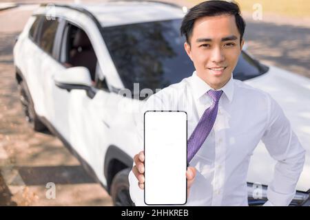 Junger lächelnder Geschäftsmann, der sein Mobiltelefon mit weißem leerem Bildschirm zeigt und vor dem Auto steht Stockfoto