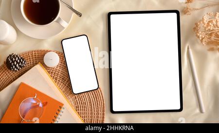 Gemütlicher, minimalistischer, femininer Arbeitsplatz mit Tablet-Computer und Smartphone-Modell mit weißem Bildschirm, Notebooks, Kaffeetasse und Zubehör. Draufsicht Stockfoto