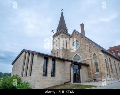 Eingang einer christlichen Backsteinkirche mit Kirchturm, Kreuz und Buntglasfenstern an einem bewölkten Abend. Stockfoto