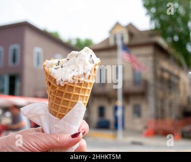 Schmelzendes Eis aus Vanille und Schokoladensplittern in Waffelkegel, teilweise gegessen, von einer Frau in der Hand gehalten, mit verschwommenem Hintergrund. Stockfoto