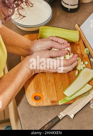 Hände der Frau schneiden mit dem Messer in Würfel oder Scheiben von jungen Zucchini Gurke auf Holz Schneidebrett. Vorbereitung von Zutaten und Gemüse vor Stockfoto