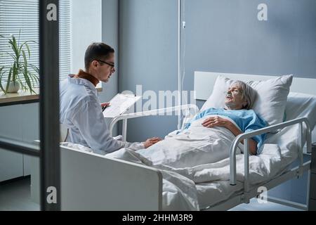 Männlicher Arzt im weißen Mantel, der eine Krankenkarte liest und mit einer kranken älteren Frau im Bett auf der Station spricht Stockfoto