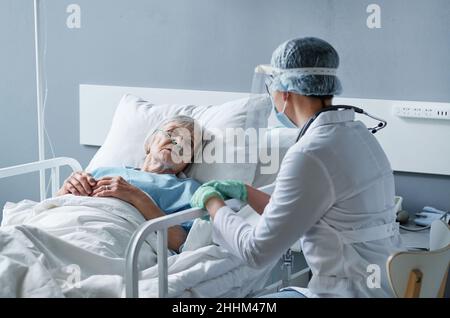 Ältere kranke Frau, die auf dem Bett liegt und mit der Ärztin in Schutzkleidung spricht, sie liegt während einer Pandemie im Krankenhaus Stockfoto