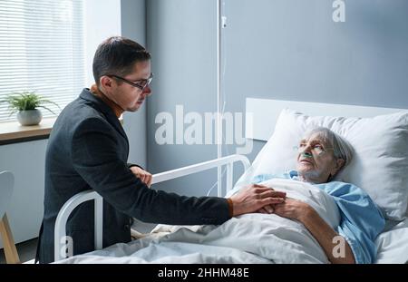 Der junge Sohn unterstützt seine ältere kranke Mutter, während sie auf der Krankenstation auf dem Bett liegt Stockfoto