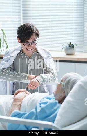 Lächelnde Tochter, die mit ihrer kranken älteren Mutter spricht, während sie während ihres Besuchs auf der Krankenhausstation auf dem Bett liegt Stockfoto