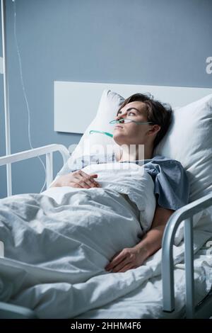 Junge Frau, die im Krankenhaus behandelt wird, sie liegt mit Sauerstoffschlauch auf dem Bett und blickt mit nachdenklichen Augen davon Stockfoto