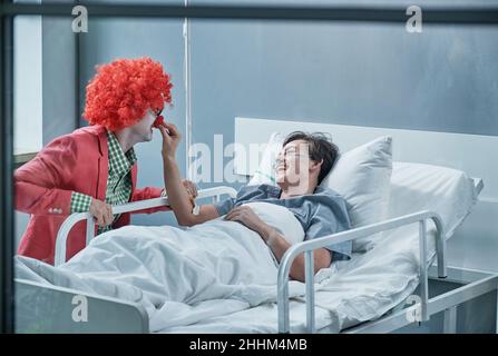 Junger Mann, der sich in einem Clownskostüm verkleidet und die kranke Frau auf der Krankenstation unterhält Stockfoto