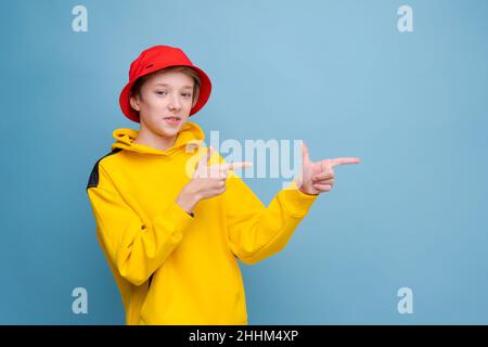 Zeigen Sie mit dem Finger zur Seite. Rapper. Informationsbanner. Stilvoller Hipster-Typ in gelbem Sweatshirt und rotem panama-Hut, der mit dem Finger auf den Kopierraum vor blauem Hintergrund zeigt Stockfoto