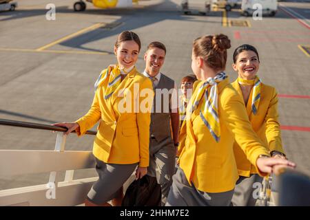 Lächelnde Männer und Frauen, die Flugbegleiter tragen, tragen Fluguniform, während sie die Flugzeugtreppe erklimmen Stockfoto