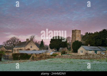 Blick über Hampnet Dorf im Winter Frost in der Morgendämmerung mit dem Vollmond und rosa Wolken am Himmel. Hampnet, Cotswolds, Gloucestershire, Großbritannien Stockfoto